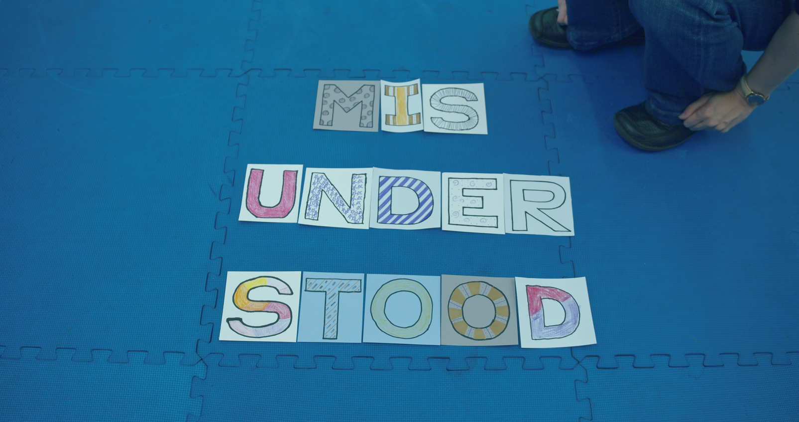 Colorful letters spelling “MISUNDERSTOOD” arranged on a blue interlocking mat, with a person kneeling nearby.