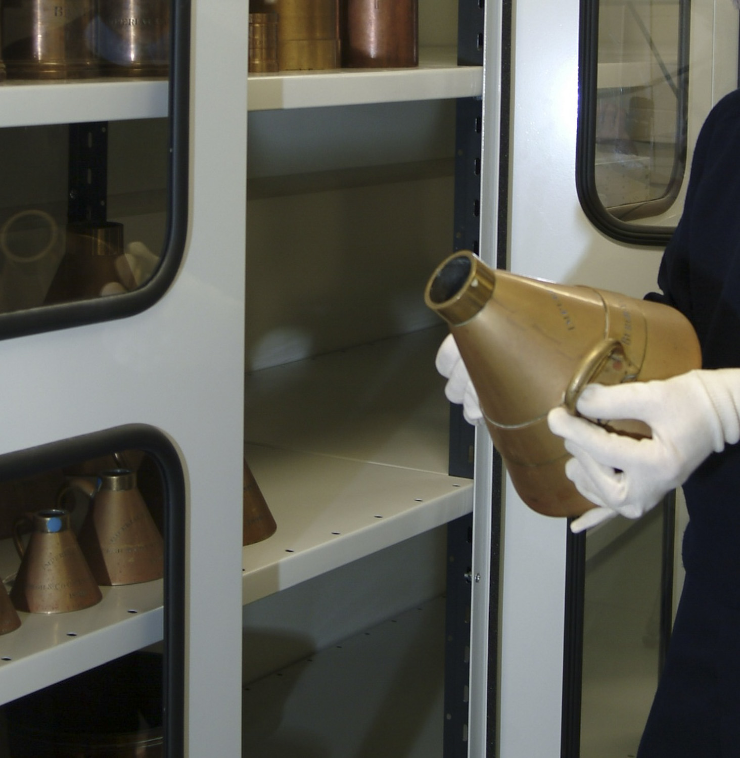 Someones gloved hands holding a brass measure. Shelves in the background.