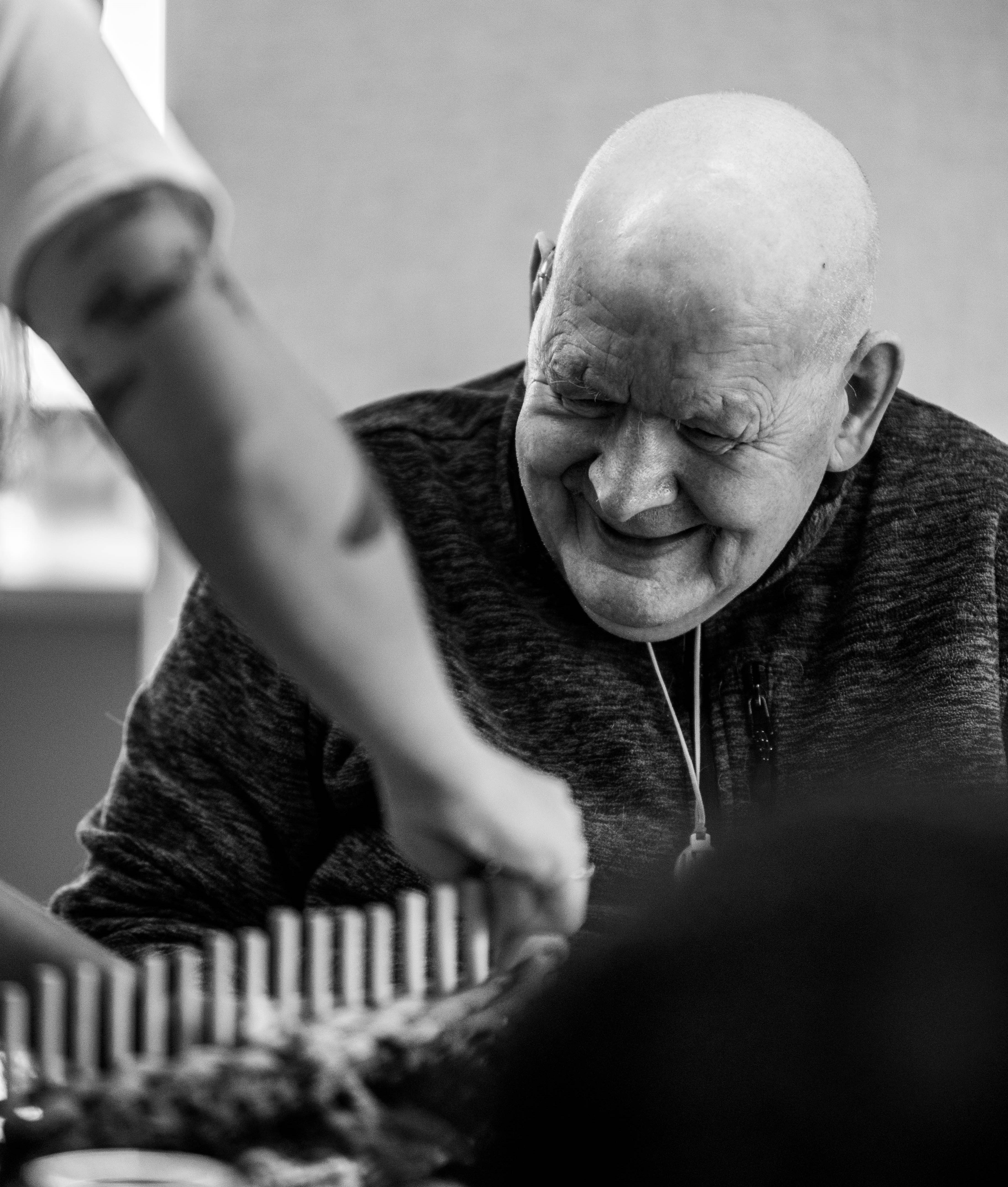 image is of an older man with no hair smiling whilst watching in the foreground the hands of the artist showing him how to weave