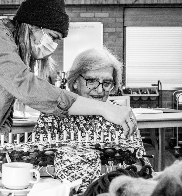 image is in black and white and of an older lady wearing a mask using her hands to feel different types of wool on a table she is sitting at