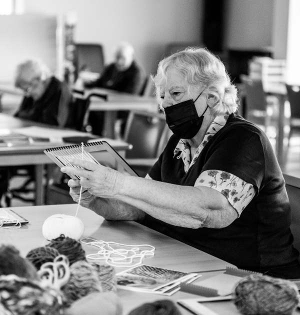 image is in black and white and of an older lady wearing a mask using her hands to feel different types of wool on a table she is sitting at