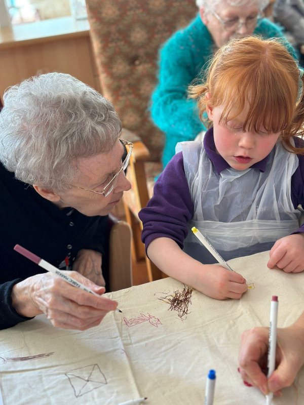 An elderly resident and young child chat together as they draw pictures